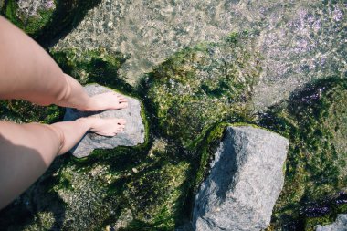 Kayanın üzerinde mavi gökyüzü ve deniz olan kadın ayakları. Deniz. Su sıçrıyor. Kıyıya. Dalgalar. Deniz köpüğü. Denizin ortasında taş var. Kum. Deniz yosunu. Yaz tatili. Deniz kıyısında bir tatil köyü. Dalgalar. Deniz yosunlu taşlar.