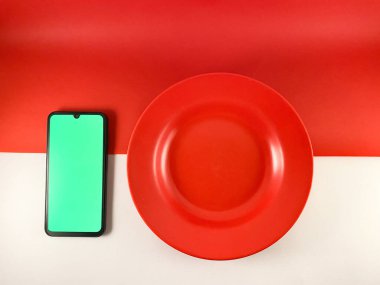 Kırmızı bir plaka, yeşil ekranlı bir telefonun sağında, sağlam kırmızı ve beyaz bir arkaplanda oturur. Tabak yuvarlaktır ve hafif çukurluk gibi görünür. Endonezya bayrak arkaplanı