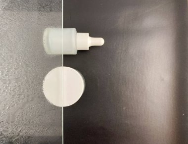 Beyaz damlalık şişesi ve beyaz krema kavanozunun beyaz ve gri eğimli arka planda yer aldığı minimalist bir kompozisyon. Cilt bakımı ürünleri, güzellik ve sağlık kavramları için mükemmel..