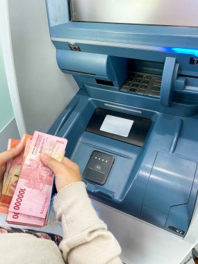 ATM makinesinin yakın plan fotoğrafı. ATM 'de gri metal bir kaplama var. Klavye, kart yuvası, ekran, makbuz makinesi ve para makinesi var. Bankamatiğin önünde para sayan biri.
