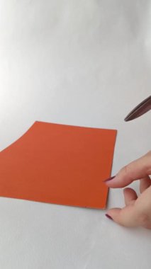 Bir el, turuncu renkli bir kağıdı makasla keser. Bir zanaat yaratma süreci, uygulama. Arka plan, mesaj için yer. Dikey, yavaş çekim videosu.