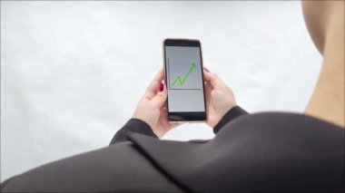 Takım elbiseli bir kadın elinde bir akıllı telefon tutuyor, ekranında da yukarı doğru büyüyen bir grafik, arkadan görülen bir görüntü. Gelir artışı. Yatırım. Yükselen fiyatlar. Kârlılık.