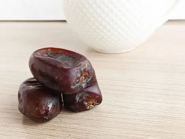 Ahşap bir masada kurumuş hurma, arka planda beyaz bir fincan sıcak çay. Çay için diyet tatlılar. Tatlı kurumuş meyveler..