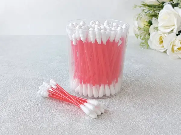 Bir yığın pamuklu çubuk ve bir kutu pamuklu bez masanın üzerinde, arka planda çiçekler var. Kulak temizleme ürünleri.