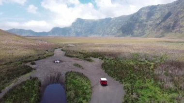 Yeşil bozkırdan geçerek dağ zirvesine doğru giden kırmızı bir off-road aracının havadan görüntüsü. Offroad aracıyla keşif..