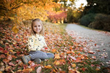 Küçük kız sonbahar parkında bankta oturuyor ve kameraya bakıyor.