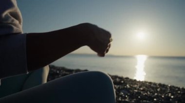 Vücut kıyafetli genç beyaz kadın sakin denizin yanındaki sahilde yoga yaparak dinleniyor. Yakın çekim elleri, Gyan Mudra ve Lotus pozisyonu. Gündoğumu arka planı. Ağır çekim. Yüksek kalite 4k görüntü