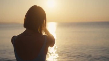 Gün batımında kadın eli cep telefonu tutuyor. Spor elbiseli genç bir kadın deniz aşırı güzel bir gün batımında cep telefonuyla fotoğraf çekiyor. Tatil sırasında akıllı telefondan fotoğraf çekmek.