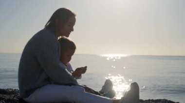 Anne deniz atında bebekle oynuyor, güzel bir gün doğumu, küçük çocuğu kucaklayan bir kadın, anne-baba sevgisi, denizin üzerinde parlayan güneş. Yüksek kalite 4k görüntü