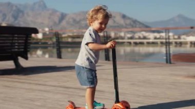 Parkta scooter kullanan küçük bir çocuk. Anaokulu çocuğu paten kaymayı öğreniyor. Güneşli yaz gününde gülümseyen çocuk paten kayıyor.