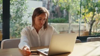Kredi kartı tutan ve dizüstü bilgisayar kullanan genç bir kadın. Evde çalışan bir iş kadını. Çevrimiçi alışveriş, e-ticaret, internet bankacılığı, harcama, ev konseptinden çalışma