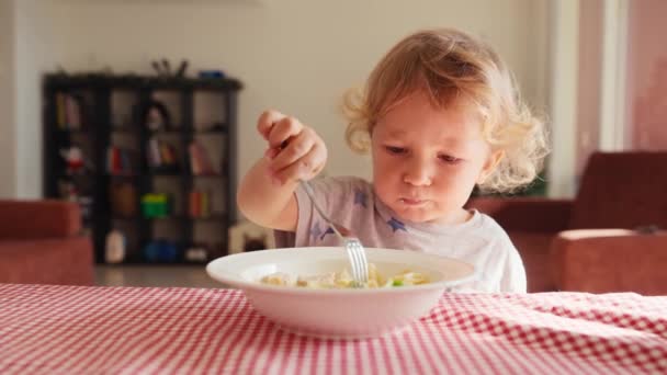 2歳の笑顔の男の子は 椅子に座って ボウルからフォークでケチャップでパスタを食べています 背景がぼやけている 子供のための健康的な食事 子供の栄養 — ストック動画