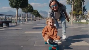 Young mother teaches ride skateboard, son of little boy, summer day. Autumnal clothes, casual clothes. Help and support parenting. Mother rides a little child on a skateboard.