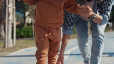 Young caucasian mother playing with her son pushing forward boy riding on skateboard in public road. Kids and entertainment. Childhood and sports concept. High quality 4k footage