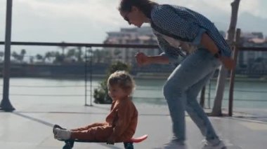 Young mother teaches ride skateboard, son of little boy, summer day. Autumnal clothes, casual clothes. Help and support parenting. Mother rides a little child on a skateboard.