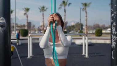 Slim young woman training pull ups with resistance band in outdoors at sunny summer day. Be H3althy