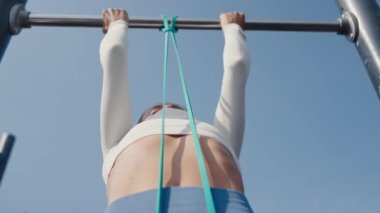 Slim young woman training pull ups with resistance band in outdoors at sunny summer day. Be H3althy
