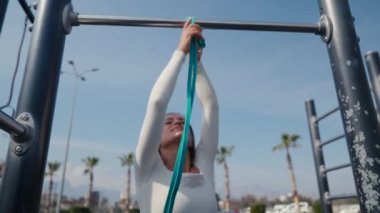 Slim young woman training pull ups with resistance band in outdoors at sunny summer day. Be H3althy