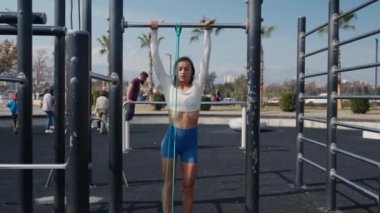 Slim young woman training pull ups with resistance band in outdoors at sunny summer day. Be H3althy