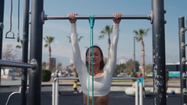 Slim young woman training pull ups with resistance band in outdoors at sunny summer day. Be H3althy