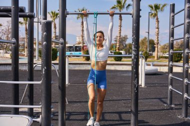 Aktif yaşam tarzı. Güneşli yaz gününde açık havada spor salonunda direniş bandıyla barfiks çeken zayıf bir genç kadın. Sağlıklı ol. Yüksek kalite fotoğraf