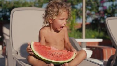 Havuzun kenarında oturup karpuz yiyen sarı saçlı güzel çocuk, yakın plan portresi. Mutlu 2 yaşındaki çocuk sulu olgun karpuz yiyor. Yüksek kalite 4k görüntü