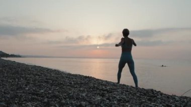 Gün batımında deniz kıyısında koşan spor elbiseli genç bir kadın. Yavaş çekim. Şortlu ve tanklı kız. Mutluluk ve özgürlük kavramı. Yüksek kalite 4k görüntü