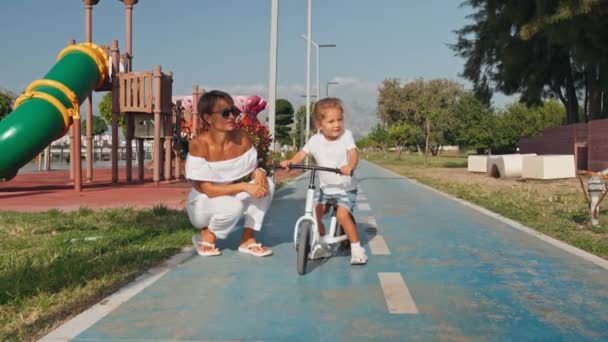 若い母親は 市立公園の白いバランスバイクで幼児の息子を捕まえる ソンライディングバイクは 都市公園の舗装に女性の親と追いつきます 高品質の4K映像 — ストック動画
