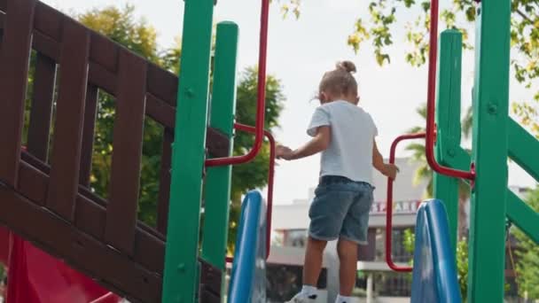 子供は暑い夏の日に公園でグリッドを登ります 多階建ての建物と近代的な住宅複合体の近代的な遊び場の小さな子供のためのロープのスライド — ストック動画