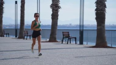 Sıska sarışın kadın gezinti güvertesinde koşuyor, şehir binaları arka planda. Sportif seksi kadın koşuyor, gün batımında dışarıda egzersiz yapıyor. Aktif yaşam tarzı, arka plan. Yüksek kalite 4k görüntü