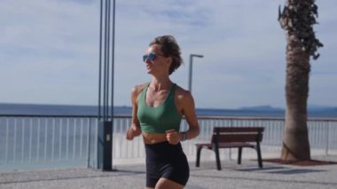 Sıska sarışın kadın gezinti güvertesinde koşuyor, şehir binaları arka planda. Sportif seksi kadın koşuyor, gün batımında dışarıda egzersiz yapıyor. Aktif yaşam tarzı, arka plan. Yüksek kalite 4k görüntü