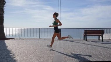 Sıska sarışın kadın gezinti güvertesinde koşuyor, şehir binaları arka planda. Sportif seksi kadın koşuyor, gün batımında dışarıda egzersiz yapıyor. Aktif yaşam tarzı, arka plan. Yüksek kalite 4k görüntü