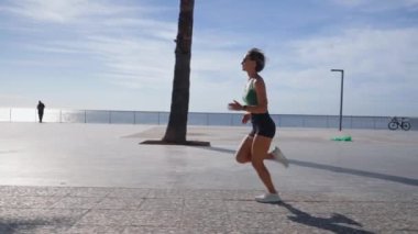 Sıska sarışın kadın gezinti güvertesinde koşuyor, şehir binaları arka planda. Sportif seksi kadın koşuyor, gün batımında dışarıda egzersiz yapıyor. Aktif yaşam tarzı, arka plan. Yüksek kalite 4k görüntü
