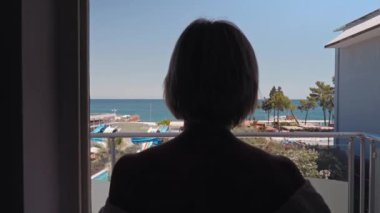 Kadın perdeleri açar ve sabah güzel deniz manzarasına hayran olmak için balkona çıkar. Tatil, Seyahat, Yaz Tatili Turizmi, Deniz Manzaralı Lüks Tatil Köyünde Dinlenme. 