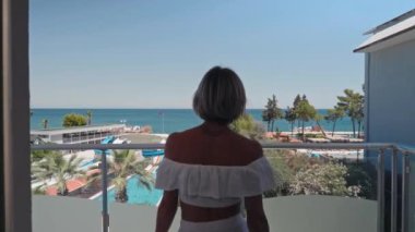Kadın perdeleri açar ve sabah güzel deniz manzarasına hayran olmak için balkona çıkar. Tatil, Seyahat, Yaz Tatili Turizmi, Deniz Manzaralı Lüks Tatil Köyünde Dinlenme. 