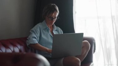 Evdeki Dizüstü bilgisayarını kullanarak şık bir oturma odasında güzel beyaz bir kadının portresi. İnterneti tarıyor ve sosyal ağlarda videoları kontrol ediyor ve eğleniyor.