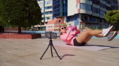 Genç bir kadın güneşli bir günde halka açık bir parkta online antrenman yapıyor. Yoga minderinde antrenman yaparken akıllı telefonuyla egzersiz talimatları izliyor. Dijital uygunluk ve kendi kendini yönlendirme kavramı