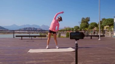 Bayan fitness eğitmeni, halka açık bir parkta akıllı telefon kamerası kullanarak idmandan önce ısınma videosu kaydeder. Güneşli bir günde dışarıda yoga minderinin üzerinde hazırlanıyor. Geniş açılı sabit açı. 