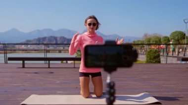 Bayan fitness eğitmeni, parktaki akıllı telefon kamerasından bir egzersiz videosu kaydediyor. Güneşli bir günde dışarıda, yoga minderinin üzerinde diz çöküyor ve arkasında dağlar var. Geniş açı statik