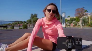 Parlak pembe spor giyimli genç atletik kadın güneşli bir günde halka açık bir parkta yoga minderinde online antrenmana başlıyor. Eğitime başlamak için kablosuz kulaklık takıyor. Bağlı açık hava kavramı