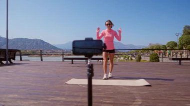 Bayan fitness eğitmeni, halka açık bir parkta akıllı telefon kamerası kullanarak idmandan önce ısınma videosu kaydeder. Güneşli bir günde dışarıda yoga minderinin üzerinde hazırlanıyor. Geniş açılı sabit açı. 