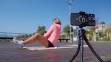 Genç bir kadın güneşli bir günde halka açık bir parkta online antrenman yapıyor. Yoga minderinde antrenman yaparken akıllı telefonuyla egzersiz talimatları izliyor. Dijital uygunluk ve kendi kendini yönlendirme kavramı