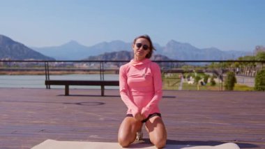 Bayan fitness eğitmeni, parktaki akıllı telefon kamerasından bir egzersiz videosu kaydediyor. Güneşli bir günde dışarıda, yoga minderinin üzerinde diz çöküyor ve arkasında dağlar var. Geniş açı statik