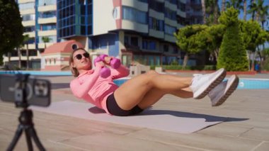 Genç bir kadın güneşli bir günde halka açık bir parkta online antrenman yapıyor. Yoga minderinde antrenman yaparken akıllı telefonuyla egzersiz talimatları izliyor. Dijital uygunluk ve kendi kendini yönlendirme kavramı