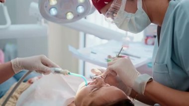 Genç bayan dişçi, modern, parlak bir diş kliniğinde bir kadın hastayı tedavi ediyor. Hasta sandalyede yatarken profesyonelce aletler kullanıyor. Kadın sağlık hizmetleri kavramı ve