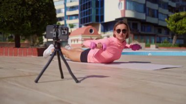 Genç bir kadın güneşli bir günde halka açık bir parkta online antrenman yapıyor. Yoga minderinde antrenman yaparken akıllı telefonuyla egzersiz talimatları izliyor. Dijital uygunluk ve kendi kendini yönlendirme kavramı