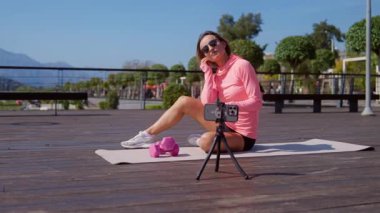 Parlak pembe spor giysili genç atletik kadın güneşli bir günde halka açık bir parkta yoga minderinde antrenman yapıyor. Akıllı telefonunu kullanarak bağımsız olarak antrenman yapıyor. Dijital uygunluk kavramı ve