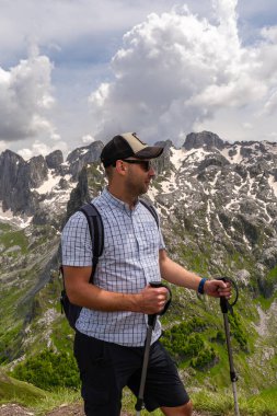 Gözlük takan, kep takan ve yürüyüş direkleri taşıyan genç bir adam karlı dağların önünde duruyor. Yeni zirveden memnun.. 