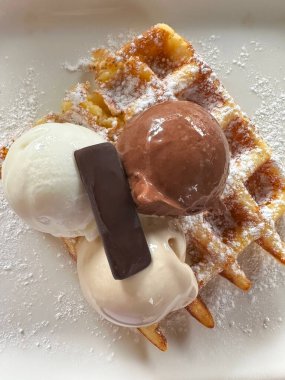 Vanilya ve çikolatalı dondurmayla kaplanmış, pudra şekeri ve bir parça çikolatayla süslenmiş ağız sulandırıcı bir waffle. Tatlı tabakta ve hazır..