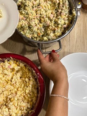Ev ortamında geleneksel Rus salatası (Olivier salatası) hazırlayan bir elin yakın çekimi. Tahta bir masada patates, yumurta ve jambon da dahil olmak üzere büyük kâseler halinde karışık malzemeler sergileniyor..